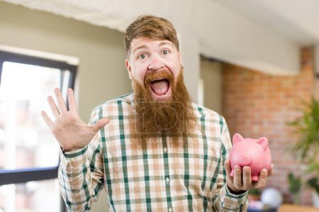 Photo for Red hair man feeling happy and astonished at something unbelievable with a piggy bank - Royalty Free Image