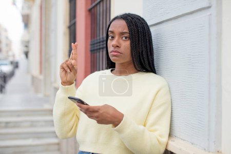 Photo for Black afro woman crossing fingers and hoping for good luck. using a smartphone concept - Royalty Free Image