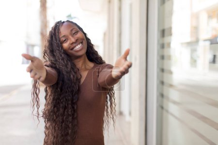Téléchargez les photos : Afro jolie femme noire souriant joyeusement donnant une accolade chaleureuse, amicale et aimante bienvenue, se sentant heureux et adorable - en image libre de droit