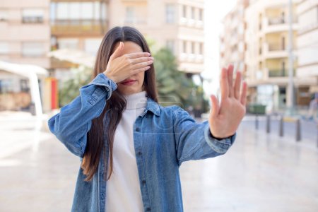 Foto de Pretty young adult woman covering face with hand and putting other hand up front to stop camera, refusing photos or pictures - Imagen libre de derechos