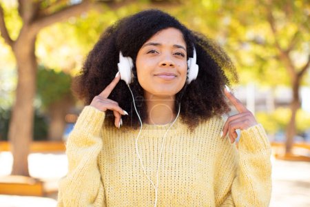 Foto de Bonita mujer negra afro sonriendo y mirando con una expresión de confianza feliz. escuchar música con auriculares - Imagen libre de derechos