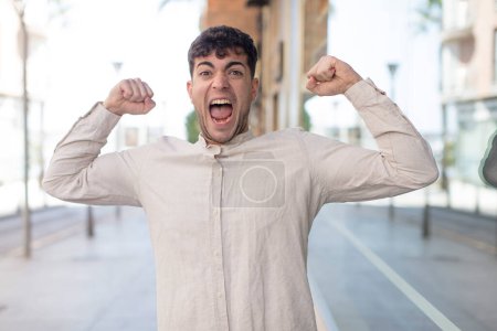 Photo for Young handsome man feeling happy, positive and successful, celebrating victory, achievements or good luck - Royalty Free Image