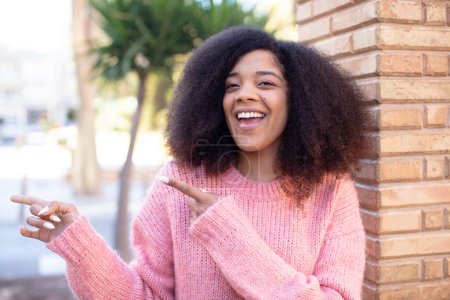 Foto de Mujer bonita afroamericana sonriendo alegremente y apuntando a copiar espacio en la palma de la mano, mostrando o publicitando un objeto - Imagen libre de derechos