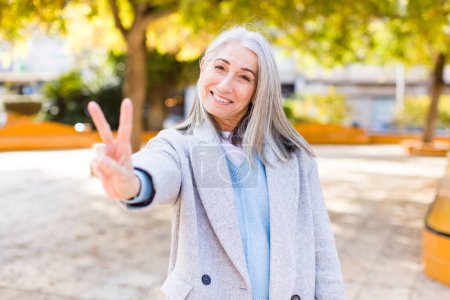 Téléchargez les photos : Senior retraité jolie femme aux cheveux blancs souriant et regardant heureux, insouciant et positif, geste de victoire ou de paix avec une main - en image libre de droit