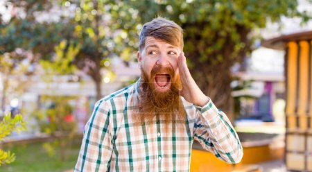 Photo for Red hair bearded man profile view, looking happy and excited, shouting and calling to copy space on the side - Royalty Free Image