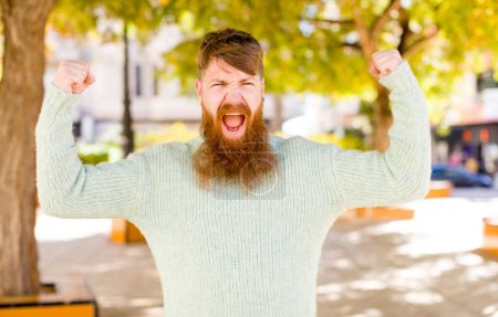 Foto de Hombre barbudo de pelo rojo gritando agresivamente con una expresión enojada o con los puños apretados celebrando el éxito - Imagen libre de derechos