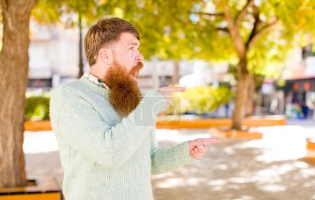 Téléchargez les photos : Cheveux roux homme barbu se sentant choqué et surpris, pointant vers copier l'espace sur le côté avec étonné, regard ouvert - en image libre de droit