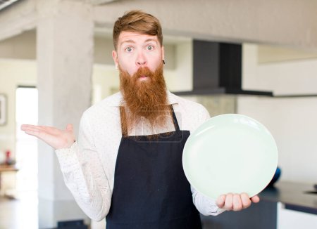 Foto de Hombre de pelo rojo encogiéndose de hombros, sintiéndose confundido e incierto con un plato vacío. concepto de chef - Imagen libre de derechos