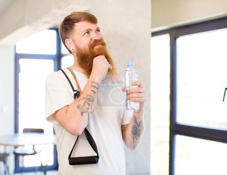 Téléchargez les photos : Homme aux cheveux roux souriant avec une expression heureuse et confiante avec la main sur le menton avec une bouteille d'eau. concept de fitness - en image libre de droit