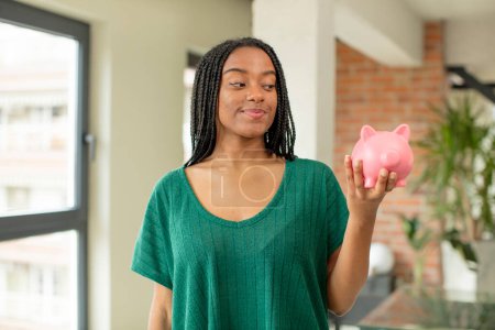 Photo for Black afro woman smiling and looking with a happy confident expression. piggy bank concept - Royalty Free Image