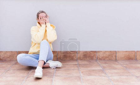 Photo for Pretty caucasian sitting  woman covering face with hands, peeking between fingers with surprised expression and looking to the side - Royalty Free Image