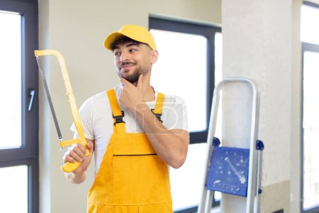 Photo for Young handsome man smiling with a happy, confident expression with hand on chin. handyman with a saw - Royalty Free Image