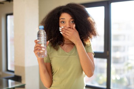 Téléchargez les photos : Jolie afro femme noire couvrant la bouche avec une main et une expression choquée ou surprise. concept de bouteille d'eau - en image libre de droit