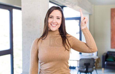 Photo for Young adult pretty woman smiling cheerfully and happily, pointing upwards with one hand to copy space - Royalty Free Image