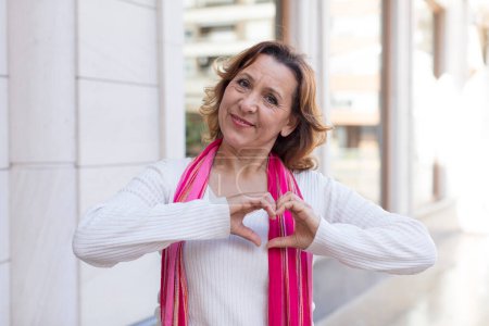 Photo for Middle age woman smiling and feeling happy, cute, romantic and in love, making heart shape with both hands - Royalty Free Image