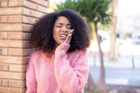 Foto de Mujer guapa afroamericana sosteniendo la mejilla y sufriendo dolor de muelas doloroso, sintiéndose mal, miserable e infeliz, buscando un dentista - Imagen libre de derechos