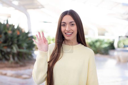 Photo for Young pretty woman smiling happily and cheerfully, waving hand, welcoming and greeting you, or saying goodbye - Royalty Free Image
