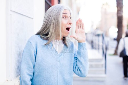 Photo for Senior retired pretty white hair woman profile view, looking happy and excited, shouting and calling to copy space on the side - Royalty Free Image