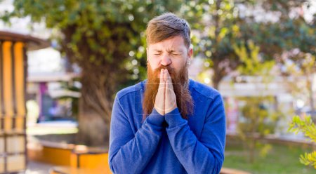 Photo for Red hair bearded man feeling worried, hopeful and religious, praying faithfully with palms pressed, begging forgiveness - Royalty Free Image