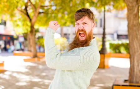 Téléchargez les photos : Cheveux roux barbu homme se sentant heureux, satisfait et puissant, ajustement flexion et biceps musculaires, regardant fort après la salle de gym - en image libre de droit