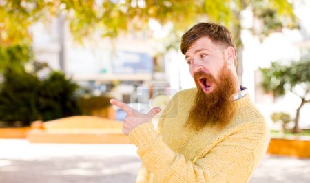 Photo for Red hair bearded man feeling joyful and surprised, smiling with a shocked expression and pointing to the side - Royalty Free Image