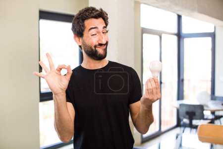 Foto de Joven barbudo loco sentirse feliz, mostrando aprobación con gesto bien con una bombilla - Imagen libre de derechos