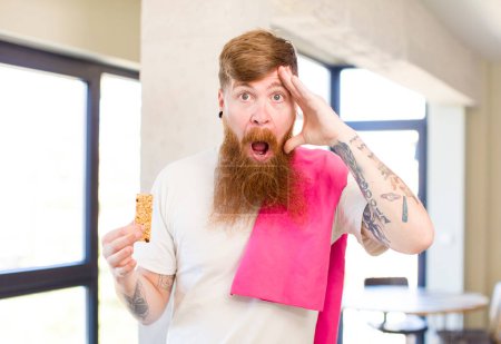 Téléchargez les photos : Homme aux cheveux roux regardant heureux, étonné et surpris avec une barre de céréales. concept de fitness - en image libre de droit