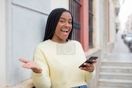 Téléchargez les photos : Afro femme noire souriant joyeusement et offrant ou montrant un concept. utilisant un concept de smartphone - en image libre de droit
