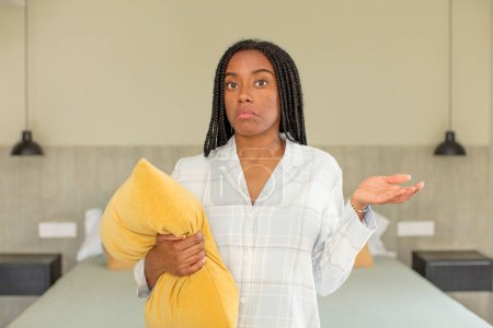 Foto de Negra afro mujer encogiéndose de hombros, sintiéndose confusa e incierta. concepto de descanso y ropa de dormir - Imagen libre de derechos