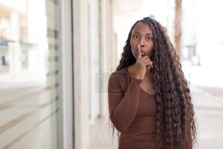 Photo for Afro pretty black woman looking serious and cross with finger pressed to lips demanding silence or quiet, keeping a secret - Royalty Free Image