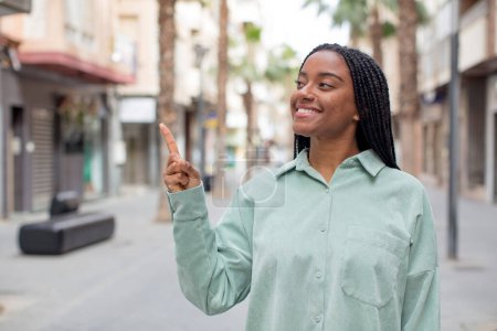 Foto de Mujer negra bonita afro sonriendo alegremente, sintiéndose feliz y señalando hacia un lado y hacia arriba, mostrando el objeto en el espacio de copia - Imagen libre de derechos