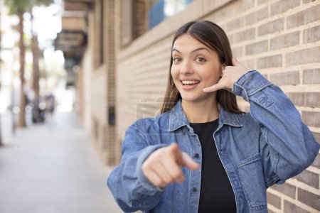 Téléchargez les photos : Jeune jolie femme souriant joyeusement et pointant vers la caméra tout en faisant un appel vous geste plus tard, parler au téléphone - en image libre de droit