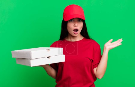 Téléchargez les photos : Hispanique jolie femme à l'air surpris et choqué, avec la mâchoire tombée tenant un objet. livraison concept de pizza - en image libre de droit