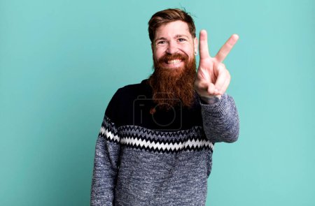 Téléchargez les photos : Longue barbe et cheveux roux homme souriant et regardant heureux, geste de victoire ou de paix - en image libre de droit