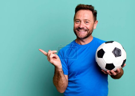 Téléchargez les photos : Homme d'âge moyen souriant joyeusement, se sentant heureux et pointant vers le côté. avec un ballon de foot. concept de fitness - en image libre de droit
