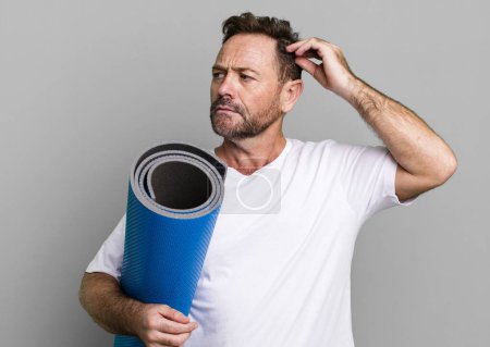 Photo for Middle age man feeling puzzled and confused, scratching head. with a yoga matt. fitness concept - Royalty Free Image