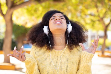 Téléchargez les photos : Jolie afro femme noire criant avec les mains en l'air. écouter de la musique avec des écouteurs - en image libre de droit