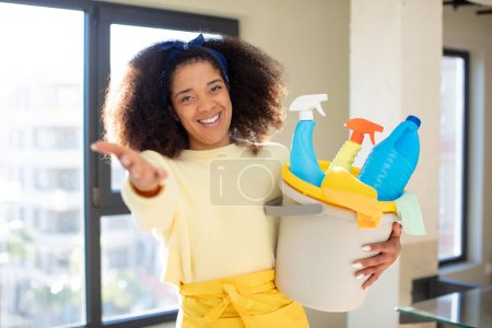 Téléchargez les photos : Jolie afro femme noire souriant joyeusement et offrant ou montrant un concept. nettoyage ménagère concept - en image libre de droit