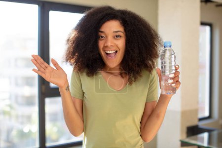 Téléchargez les photos : Jolie afro femme noire se sentant heureuse et étonnée de quelque chose d'incroyable. concept de bouteille d'eau - en image libre de droit
