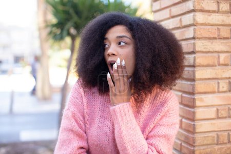 Téléchargez les photos : Afro-américaine jolie femme se sentant choquée et étonnée tenant face à face dans l'incrédulité avec la bouche grande ouverte - en image libre de droit