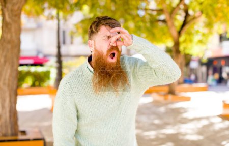 Téléchargez les photos : Cheveux roux homme barbu se sentant dégoûté, tenant le nez pour éviter de sentir une puanteur sale et désagréable - en image libre de droit