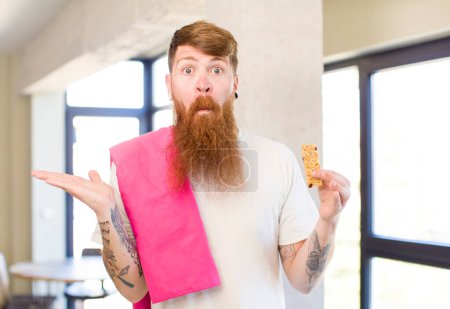 Foto de Hombre de pelo rojo encogiéndose de hombros, sintiéndose confundido e incierto con una barra de cereales. concepto de fitness - Imagen libre de derechos
