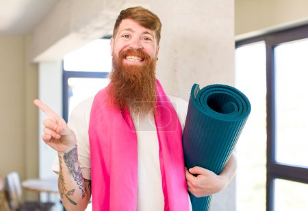 Foto de Hombre de pelo rojo sonriendo alegremente, sintiéndose feliz y señalando hacia un lado con un yoga mate. concepto de fitness - Imagen libre de derechos
