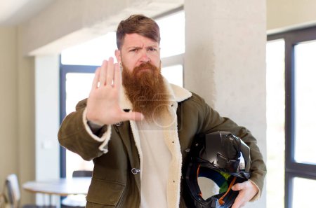 Foto de Hombre de pelo rojo mirando serio mostrando palmera abierta haciendo gesto de parada con un casco de moto - Imagen libre de derechos