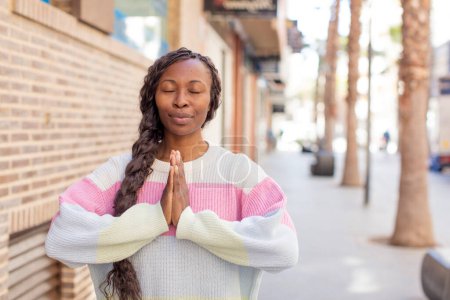 Téléchargez les photos : Afro jolie femme noire se sentant inquiète, pleine d'espoir et religieuse, priant fidèlement avec les paumes pressées, implorant pardon - en image libre de droit