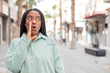 Téléchargez les photos : Afro jolie femme noire se sentant choquée et étonnée tenant face à face dans l'incrédulité avec la bouche grande ouverte - en image libre de droit