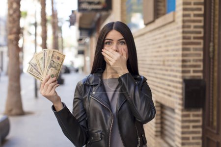 Foto de Mujer joven y bonita cubriendo la boca con una mano y expresión sorprendida o sorprendida. concepto de billetes en dólares - Imagen libre de derechos