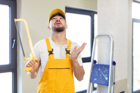 Photo for Young handsome man screaming with hands up in the air. handyman with a saw - Royalty Free Image