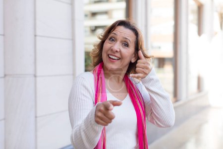 Foto de Mujer de mediana edad sonriendo alegremente y señalando a la cámara mientras que hacer una llamada más tarde gesto, hablando por teléfono - Imagen libre de derechos