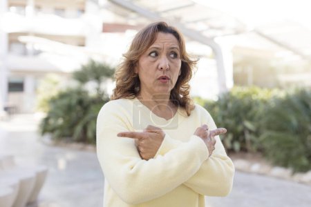 Téléchargez les photos : Femme d'âge moyen regardant perplexe et confus, précaire et pointant dans des directions opposées avec des doutes - en image libre de droit